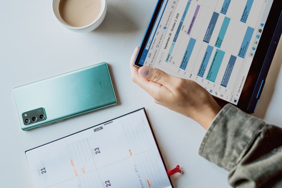 A person holding a laptop showing a calendar schedule next to a planner and phone