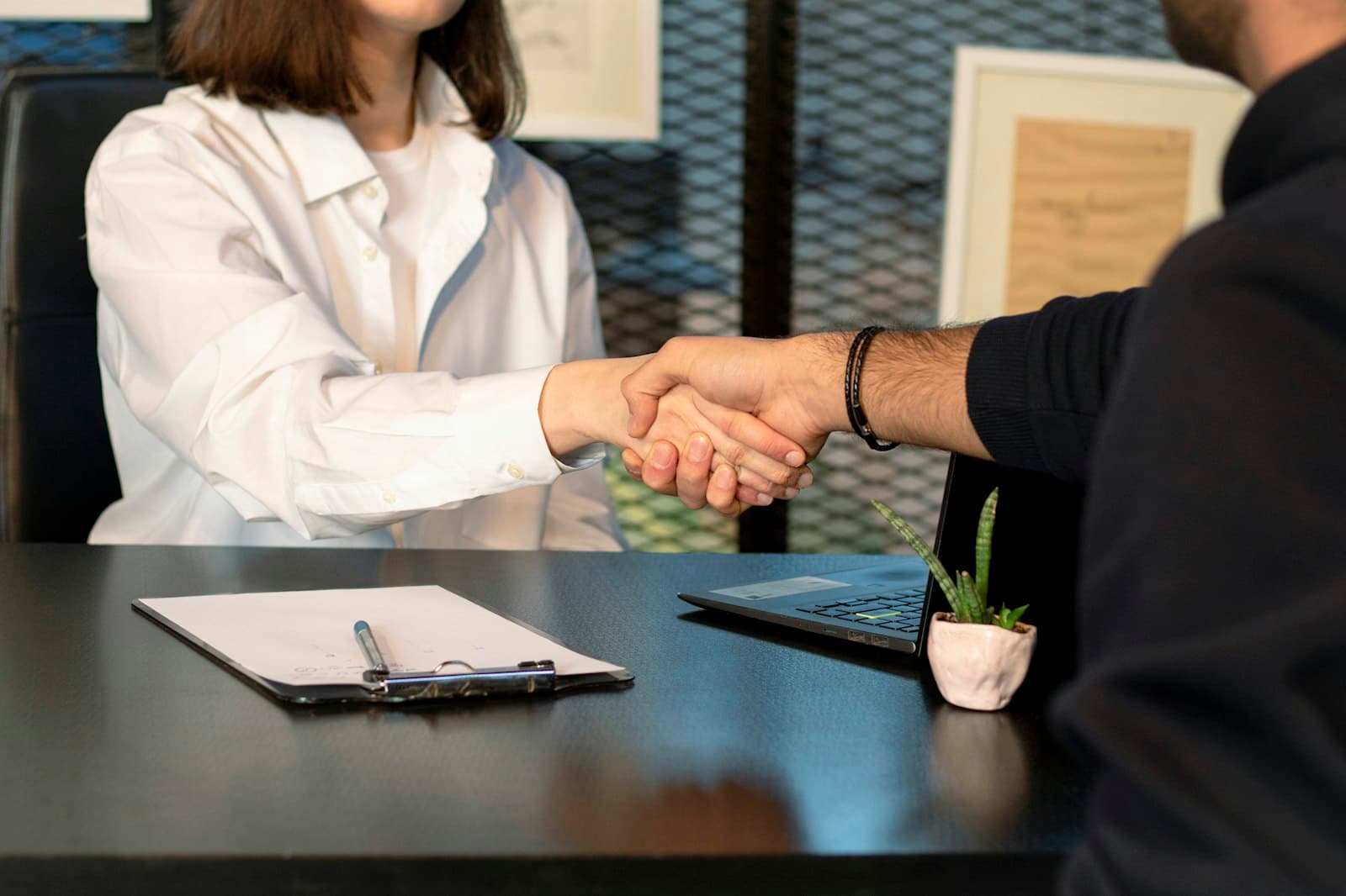 Two colleagues shaking hands across a desk with a clipboard and laptop, representing a new hire welcome